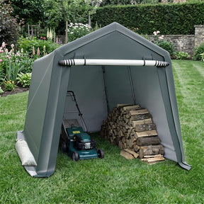 An outdoor garden shed tent in dark green on a grassy lawn, with a lawnmower and a stack of firewood stored inside. The background shows a landscaped garden with flowers and a stone wall.