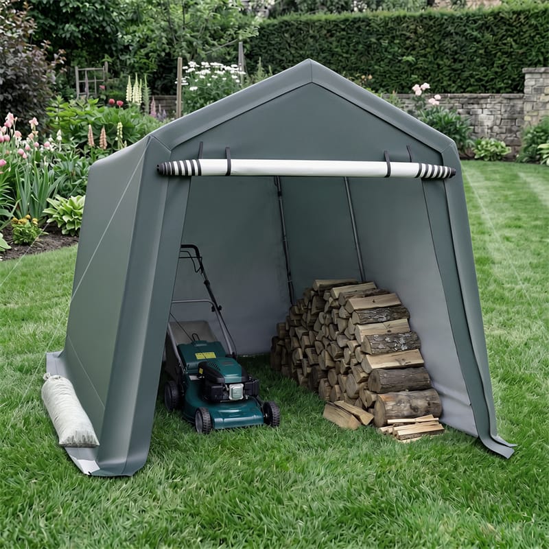 An outdoor garden shed tent in dark green on a grassy lawn, with a lawnmower and a stack of firewood stored inside. The background shows a landscaped garden with flowers and a stone wall.